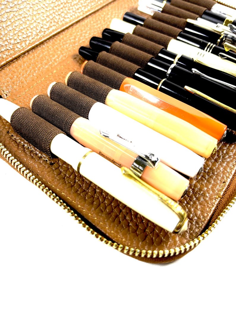 May include: A brown leather pen case with a gold zipper, filled with various fountain pens. The pens are in different colors, including black, white, orange, and cream, with gold and silver accents. The case is open, revealing the pens.