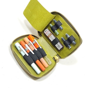 May include: Open, lime green leather pen case with a zipper closure. The case contains several pens in various colors, including orange, white, and light blue. Also included are mechanical pencils and lead refills. The case is open, revealing its contents.