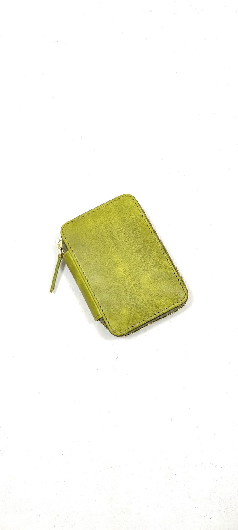 May include: A small, rectangular, olive green leather wallet with a zipper closure. The wallet has a textured surface and a gold-colored zipper. The zipper pull is made of the same leather as the wallet.
