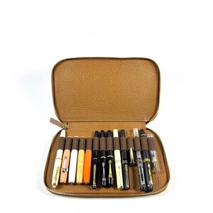May include: An open, brown leather pen case filled with a collection of fountain pens. The pens vary in color, including white, orange, black, and gold, and are arranged neatly inside the case. The case is zippered and has a textured surface.