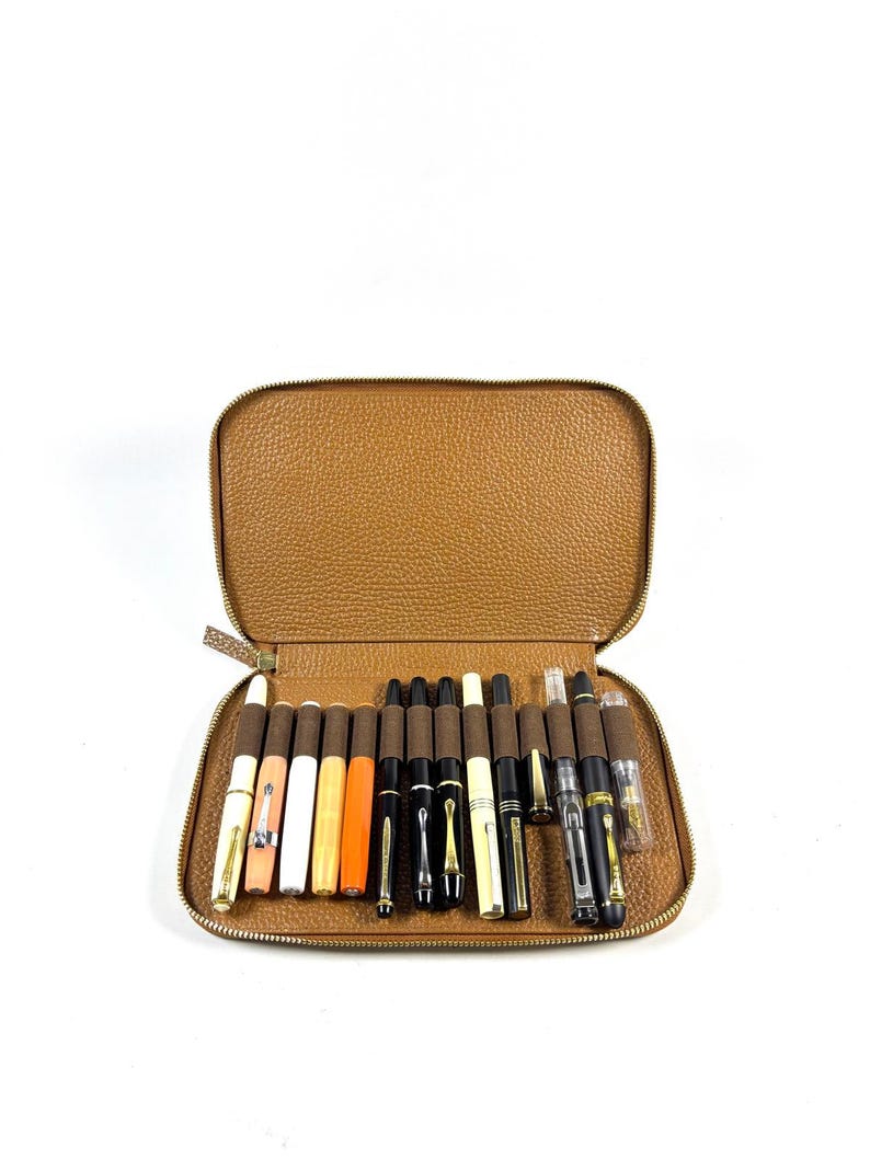 May include: An open, brown leather pen case filled with a collection of fountain pens. The pens vary in color, including white, orange, black, and gold, and are arranged neatly inside the case. The case is zippered and has a textured surface.