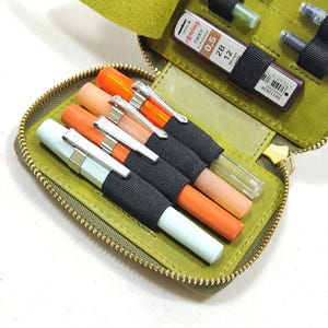 May include: Open olive green leather pen case with a gold zipper, holding several pens in various colors. The case also contains a small container of 0.5 mm lead refills.