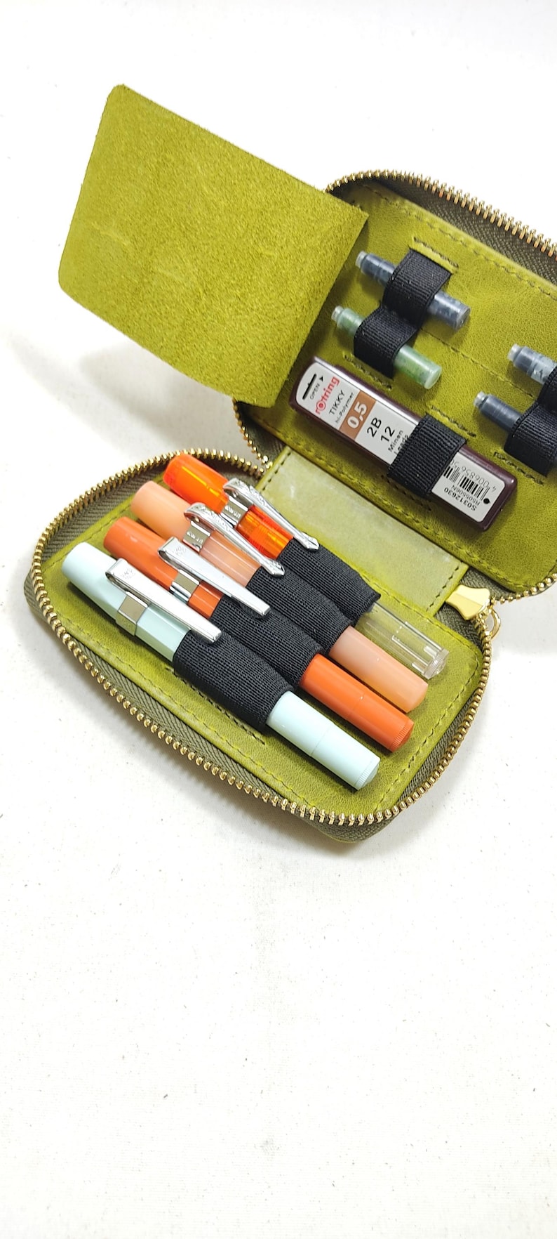 May include: Open olive green leather pen case with a gold zipper, holding several pens in various colors. The case also contains a small container of 0.5 mm lead refills.
