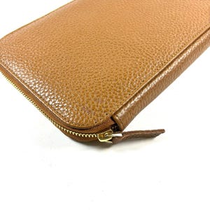 May include: A close-up of a tan leather wallet with a gold zipper. The wallet has a textured surface and a rounded shape. The zipper is partially open, revealing the interior.