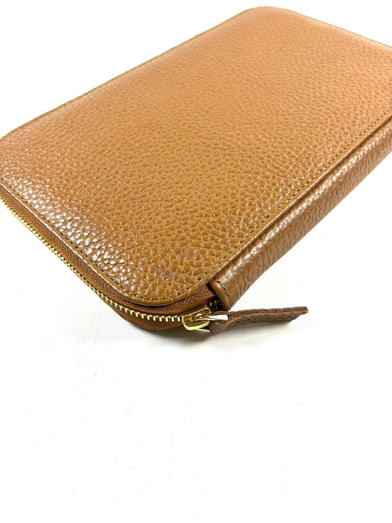May include: A close-up of a tan leather wallet with a gold zipper. The wallet has a textured surface and a rounded shape. The zipper is partially open, revealing the interior.