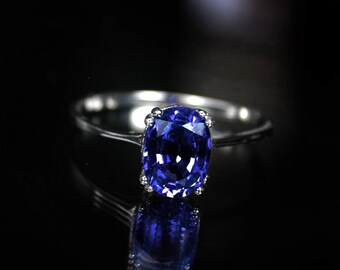 Op de afbeelding: Een zilveren ring met een grote, ovale saffier-edelsteen. De saffier is diepblauw en is gezet in een kraalbezetting. De ring ligt op een zwarte achtergrond.
