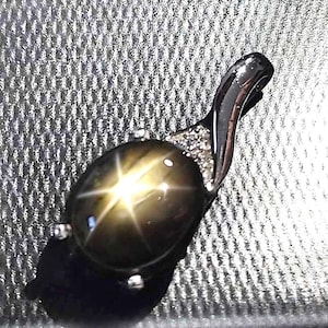 May include: A dark gemstone pendant with a star-like pattern. The oval-shaped stone is set in a silver-coloured metal setting with small, sparkling accents. The pendant has a curved, elegant design.