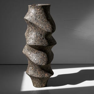 May include: A tall, dark gray ceramic vase with a unique, twisted, spiral design. The vase has a rough, textured surface.