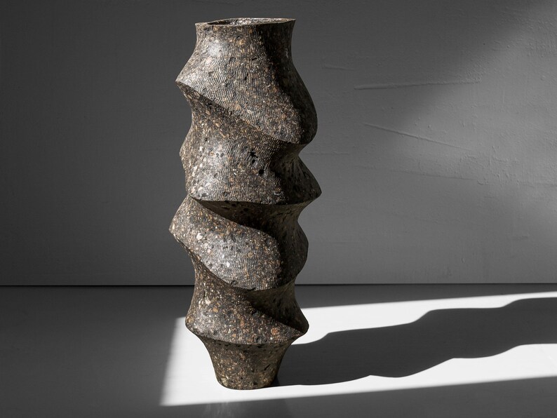 May include: A tall, dark gray ceramic vase with a unique, twisted, spiral design. The vase has a rough, textured surface.