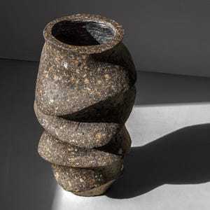 May include: A brown ceramic vase with a textured, speckled surface. The vase has a unique, twisted shape with multiple layers.