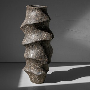 May include: A dark gray ceramic vase with a unique, twisted, and textured design. The vase is tall and narrow, with a rounded base and a slightly flared opening.