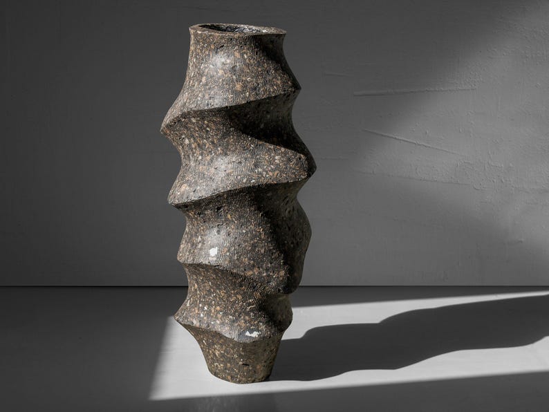May include: A dark gray ceramic vase with a unique, twisted, and textured design. The vase is tall and narrow, with a rounded base and a slightly flared opening.