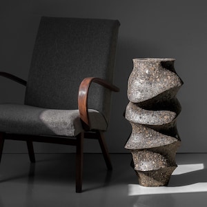May include: A gray armchair with wooden arms and a gray fabric seat sits next to a large, gray, textured, ceramic vase with a spiral design.
