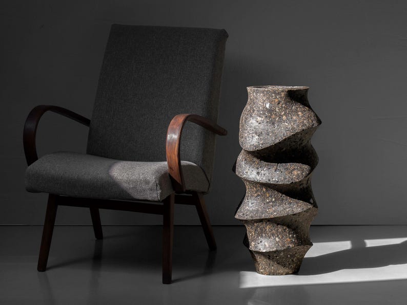 May include: A gray armchair with wooden arms and a gray fabric seat sits next to a large, gray, textured, ceramic vase with a spiral design.