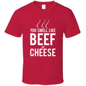 May include: A red t-shirt with white text that reads "YOU SMELL LIKE BEEF AND CHEESE". Above the text are three curved lines. The shirt is a short-sleeved crew neck.
