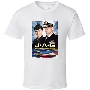 May include: White t-shirt featuring a graphic from the television show "JAG." The graphic includes two people in navy uniforms, the show's title, and a gavel over an American flag. The shirt is a classic crew neck style.