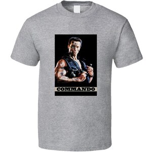 May include: A gray t-shirt featuring a rectangular graphic of a man in a tactical vest, holding a weapon. The graphic is set against a black background, with the word "COMMANDO" in white text below the image.