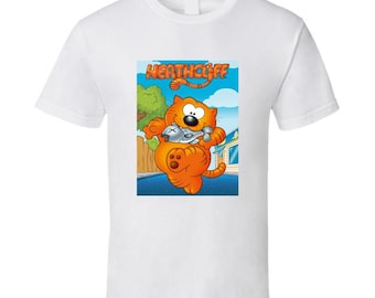 May include: White t-shirt featuring a cartoon image of Heathcliff, an orange cat holding a fish. The word "Heathcliff" is printed in orange letters above the cat. The background shows a blue sky, trees, and a house.