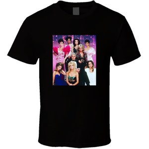 May include: A black t-shirt featuring a colourful image of the cast from the television show 'Dynasty'. The image shows eight people, including a man and seven women, all dressed in formal attire. The background is a sparkly, pink and purple design.