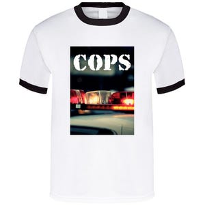 May include: White ringer t-shirt with black trim. The front displays a graphic of a police car's flashing lights and the word "COPS" in white, bold letters. The image is centrally placed on the shirt.