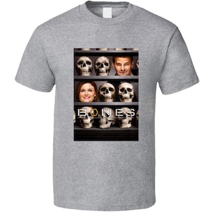 May include: A gray t-shirt featuring a graphic from the television show "Bones." The graphic includes images of skulls, actors, and the show's title. The shirt is made of a soft material.