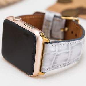 May include: A gold-colored smartwatch with a black screen and a light gray, textured leather band. The band features a gold-colored buckle and a brown leather backing. The watch is angled, showcasing its design and details.