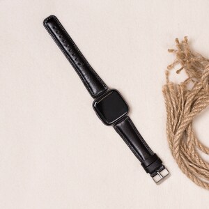 May include: A black smartwatch with a black leather band. The band has a silver buckle and is connected to the watch face. The watch is displayed on a cream-colored surface, with a bundle of brown rope in the background.