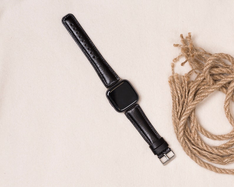 May include: A black smartwatch with a black leather band. The band has a silver buckle and is connected to the watch face. The watch is displayed on a cream-colored surface, with a bundle of brown rope in the background.