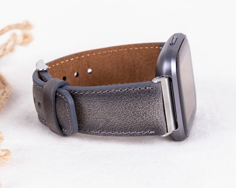 May include: A gray leather watch band with silver hardware. The band is designed for a smartwatch with a square face.