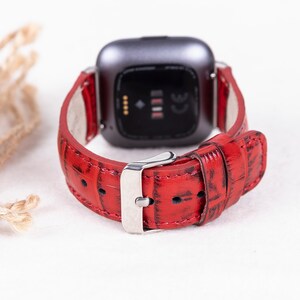 May include: Red leather watch band with a silver buckle. The band is designed for a smartwatch with a square face.