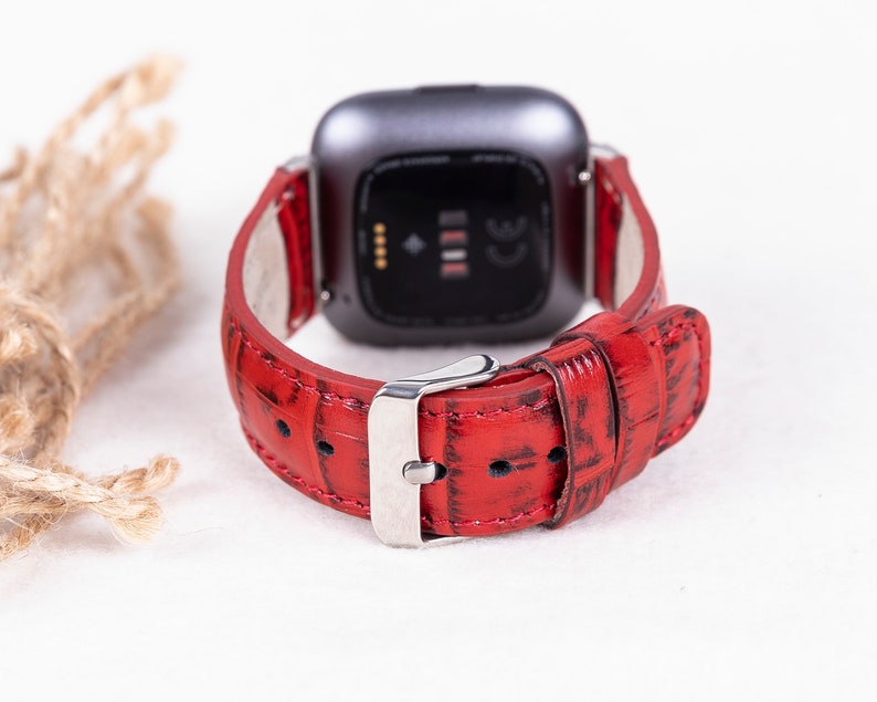 May include: Red leather watch band with a silver buckle. The band is designed for a smartwatch with a square face.