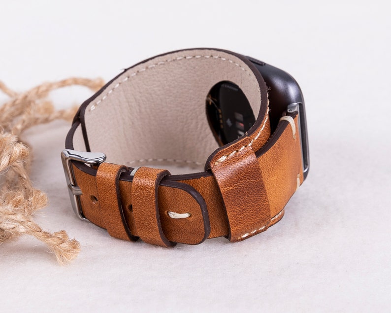 May include: A brown leather watch strap with white stitching and a silver buckle. The strap is designed for a smartwatch and has a loop to secure the strap.