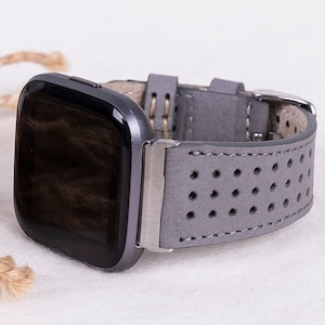 Puede incluir: Una correa de reloj de cuero gris con un dise&ntilde;o perforado. La correa est&aacute; unida a un reloj inteligente negro con una esfera cuadrada.
