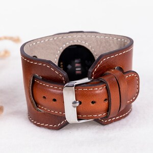 K&ouml;nnte beinhalten: Ein braunes Lederarmband mit wei&szlig;en N&auml;hten und einer silbernen Schnalle. Das Armband ist f&uuml;r eine Smartwatch konzipiert.