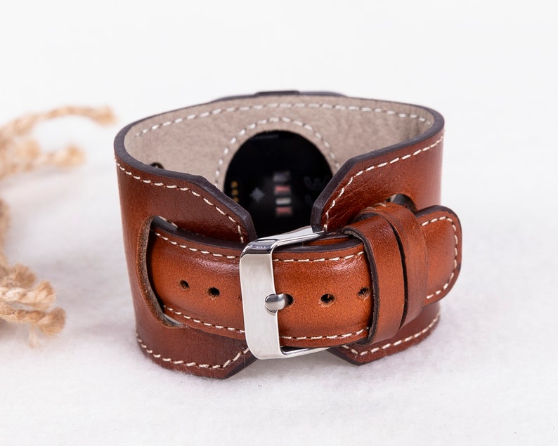 K&ouml;nnte beinhalten: Ein braunes Lederarmband mit wei&szlig;en N&auml;hten und einer silbernen Schnalle. Das Armband ist f&uuml;r eine Smartwatch konzipiert.