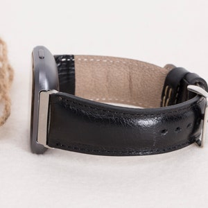 May include: A black leather watch band with a silver buckle is shown. The band is attached to a dark gray smartwatch. The watch band has a light brown interior and is stitched with white thread. The watch is displayed on a white surface.
