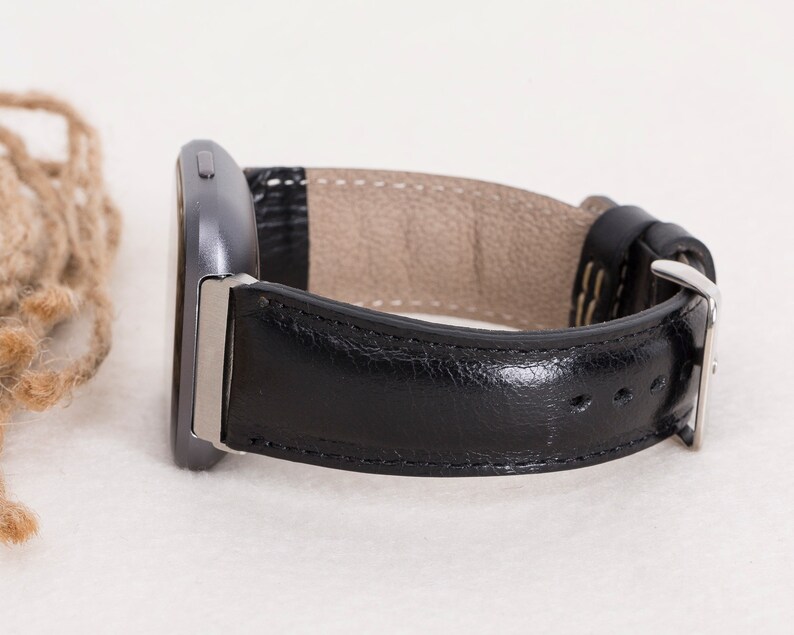 May include: A black leather watch band with a silver buckle is shown. The band is attached to a dark gray smartwatch. The watch band has a light brown interior and is stitched with white thread. The watch is displayed on a white surface.