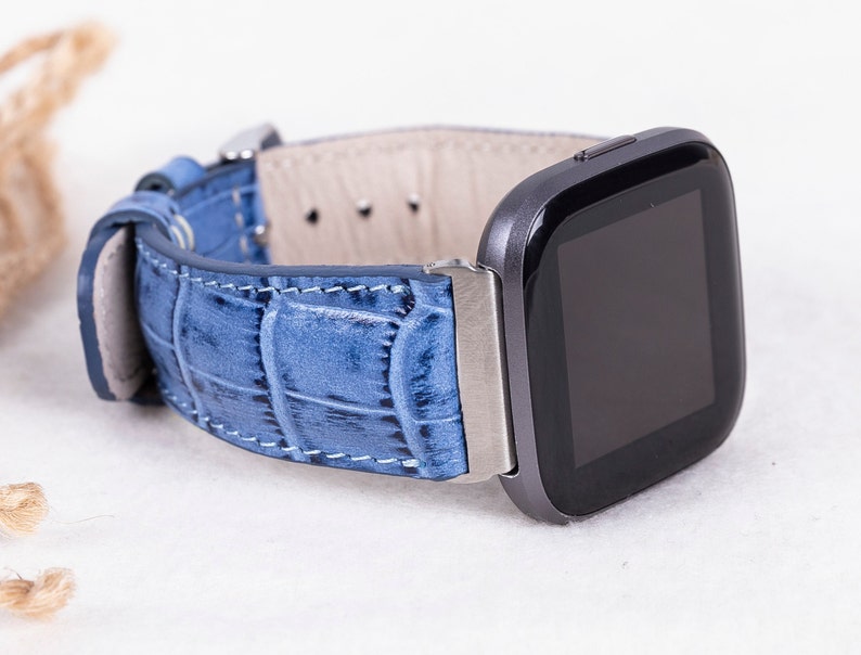 Puede incluir: Una correa de reloj de cuero azul con un patr&oacute;n de cocodrilo. La correa est&aacute; unida a un reloj inteligente negro y plateado.