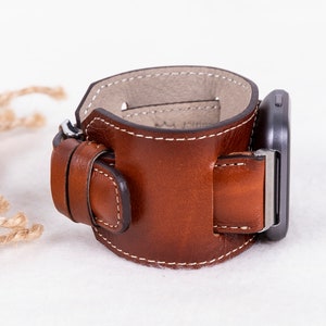 K&ouml;nnte beinhalten: Ein braunes Lederarmband mit einer Schlie&szlig;e. Das Armband ist so konzipiert, dass es sich um das Handgelenk legt und einen Riemen hat, der durch die Schlie&szlig;e geht.