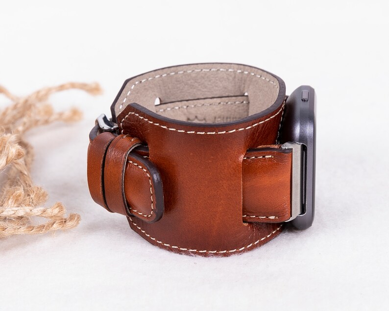 K&ouml;nnte beinhalten: Ein braunes Lederarmband mit einer Schlie&szlig;e. Das Armband ist so konzipiert, dass es sich um das Handgelenk legt und einen Riemen hat, der durch die Schlie&szlig;e geht.