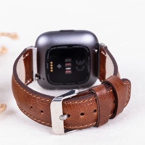 May include: A brown leather watch band with a silver buckle. The band is attached to a gray smartwatch with a black screen.
