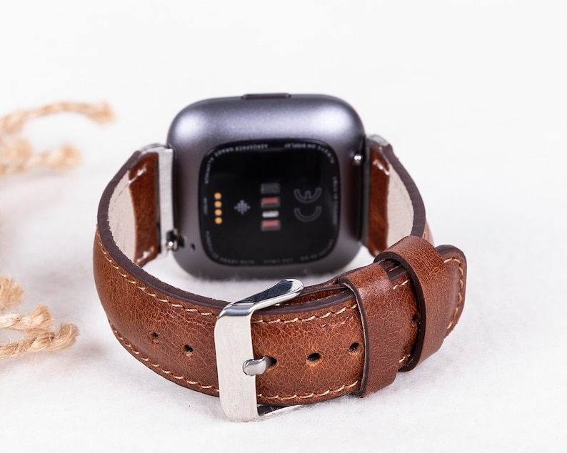 May include: A brown leather watch band with a silver buckle. The band is attached to a gray smartwatch with a black screen.
