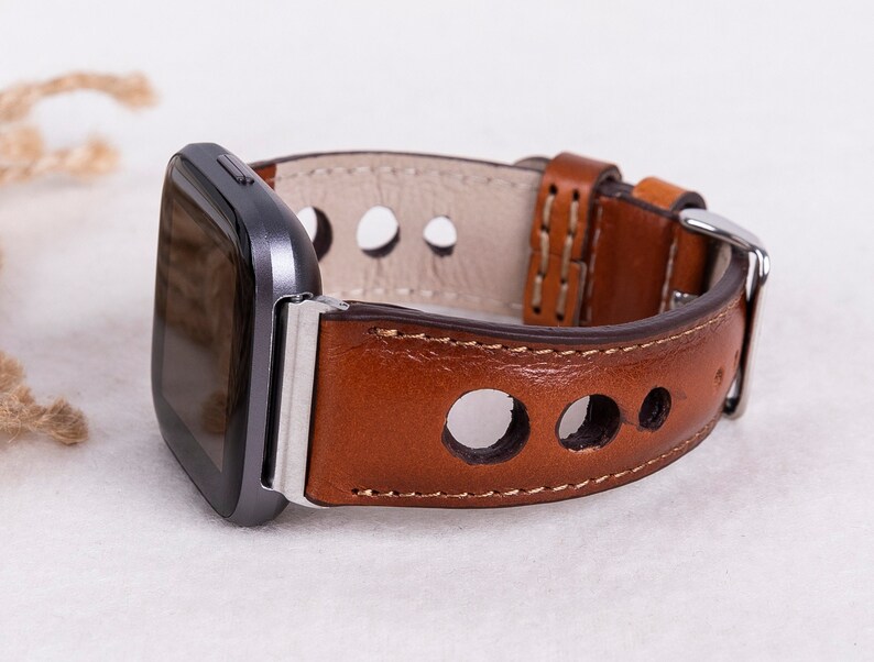 May include: A brown leather watch band with a silver buckle and a gray smartwatch. The band has a perforated design with round holes.