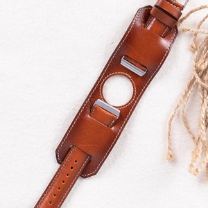 K&ouml;nnte beinhalten: Ein braunes Lederarmband mit wei&szlig;en N&auml;hten und einer silbernen Schlie&szlig;e. Das Armband hat ein einzigartiges Design mit einem Loch in der Mitte und zwei zus&auml;tzlichen B&auml;ndern, die verstellt werden k&ouml;nnen.