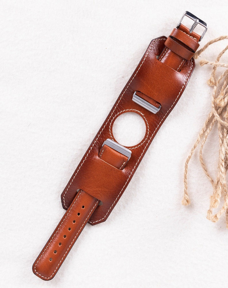 K&ouml;nnte beinhalten: Ein braunes Lederarmband mit wei&szlig;en N&auml;hten und einer silbernen Schlie&szlig;e. Das Armband hat ein einzigartiges Design mit einem Loch in der Mitte und zwei zus&auml;tzlichen B&auml;ndern, die verstellt werden k&ouml;nnen.