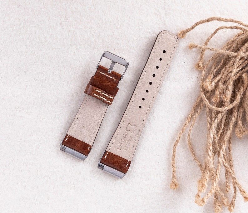 May include: Two brown leather watch straps with white stitching. The strap on the left has a silver buckle. The strap on the right has the text "Full Grain Leather" printed on it.