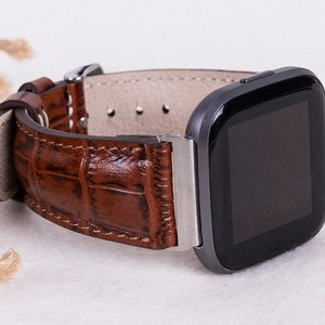 May include: A brown leather watch band with a crocodile pattern. The band is attached to a black smartwatch with a square face.