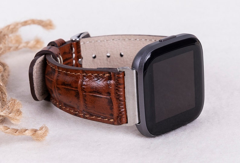 May include: A brown leather watch band with a crocodile pattern. The band is attached to a black smartwatch with a square face.