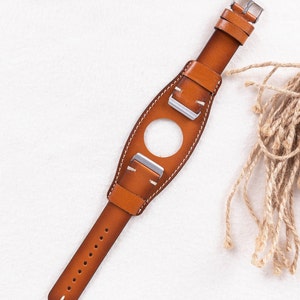 Puede incluir: Una correa de reloj de cuero marr&oacute;n con hebillas plateadas. La correa tiene un agujero en el medio para una esfera de reloj.