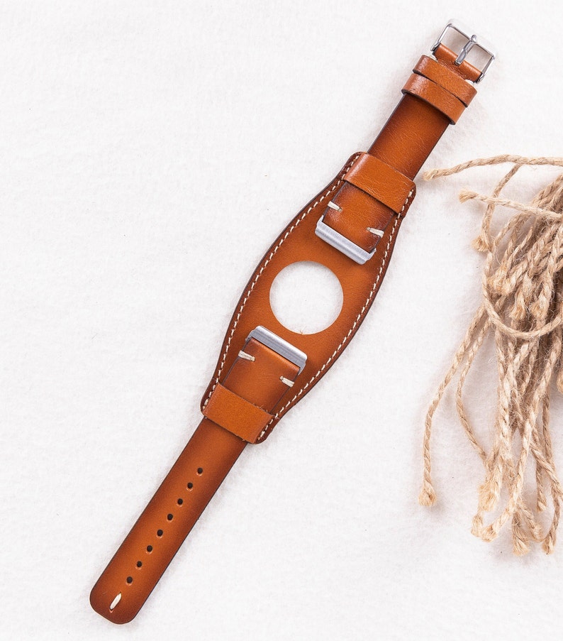Puede incluir: Una correa de reloj de cuero marr&oacute;n con hebillas plateadas. La correa tiene un agujero en el medio para una esfera de reloj.